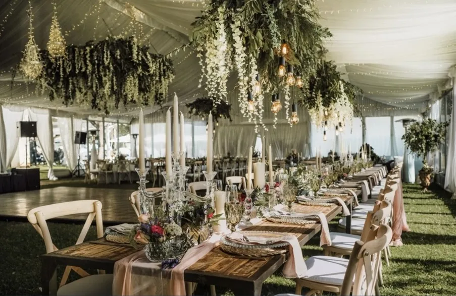 Luxurious wedding reception setup in a draped marquee tent with a long wooden table, elegant candelabras, and dramatic cascading white and green floral installations overhead.