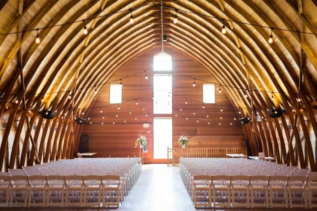 Rustic barn wedding ceremony setup with white folding chairs arranged on a wooden floor, soft string lights and a crystal chandelier hanging from exposed beams, and an open doorway overlooking green trees.
