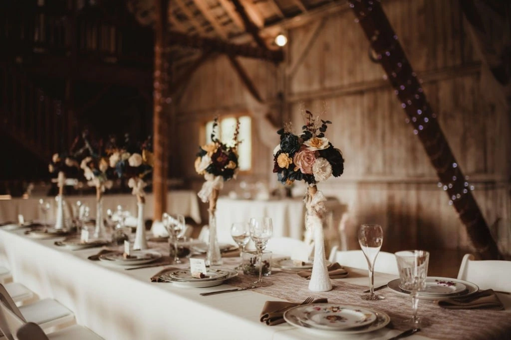 Long rustic wedding reception table set up inside a barn, featuring tall white vases with dark floral centerpieces, vintage plates, cloth napkins, and a burlap table runner.