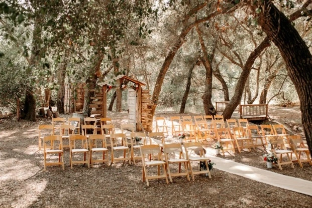 Rustic outdoor wedding ceremony setting with rows of light wooden folding chairs, a white aisle runner leading to a rustic wooden arch decorated with greenery and flowers, surrounded by tall forest trees in natural light.