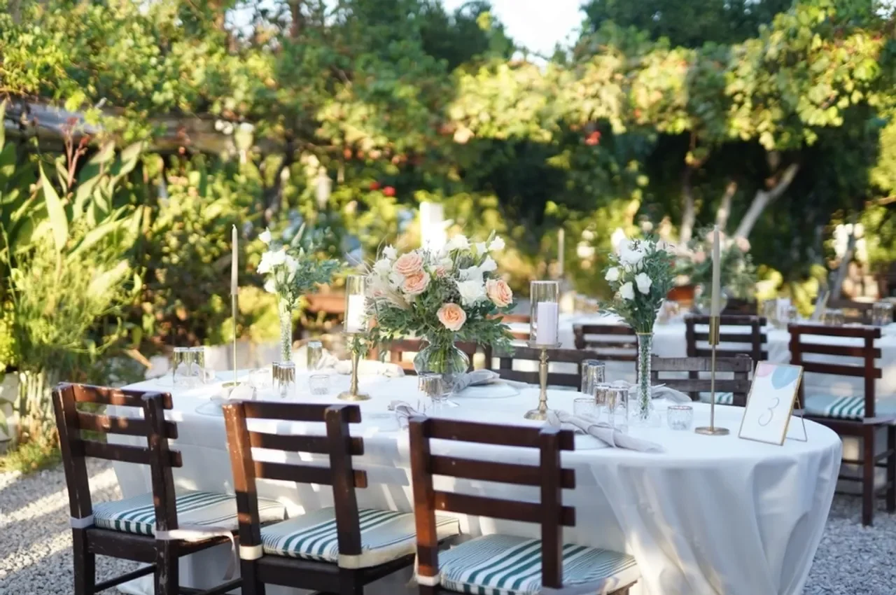Outdoor garden wedding reception table with white table linens, wooden chairs, elegant floral centerpieces in soft blush and greenery, and tall white candles set among lush trees in natural sunlight.