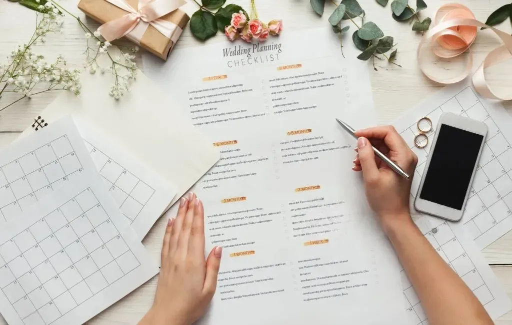Top-down view of a person’s hands using a silver pen to fill out a wedding planning checklist on a white wooden desk, surrounded by a calendar, a smartphone, wedding rings, pink roses, and a small gift box.